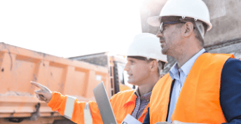 Two men in orange safety vest and jacket on a construction site talking to each other