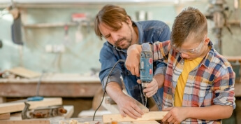 Father working in wood shop with son helping him hold a drill